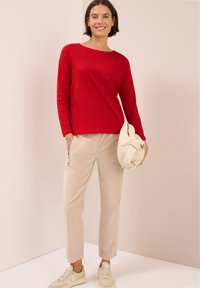 Maglietta rossa a maniche lunghe con scollatura a barca, abbinata a pantaloni beige con zip laterale e sneaker di colore chiaro.