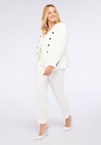 Blazer bianco con bottoni decorativi neri, abbinato a pantaloni bianchi su misura e décolleté bianchi a punta. Tessuto liscio, design strutturato.