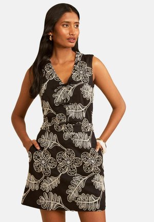 Frau trägt ein ärmelloses schwarzes Kleid mit großen weißen Blumen- und Blättermustern, Hände in den Taschen, schaut zur Seite.