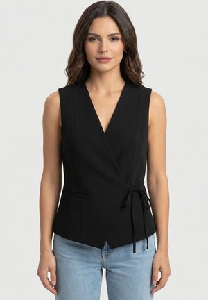 Donna con capelli castani alla lunghezza delle spalle che indossa un gilet nero senza maniche annodato in vita e jeans azzurri chiari su sfondo bianco.