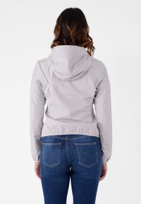 Giacca grigia chiara con zip e cappuccio, vita elasticizzata e polsini a coste, indossata sopra jeans blu con tasche posteriori curve.