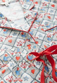 Baumwoll-Pyjama-Set mit weißem Hintergrund, das ein Patchwork-Muster aus blauen Bären, roten Herzen, Blumen und Bändern zeigt. Rote Akzentnähte.