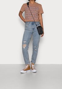Persona que lleva una camiseta gráfica marrón y jeans azules desgastados, de pie con un pequeño bolso cruzado negro y zapatillas color beige sobre un fondo neutro.