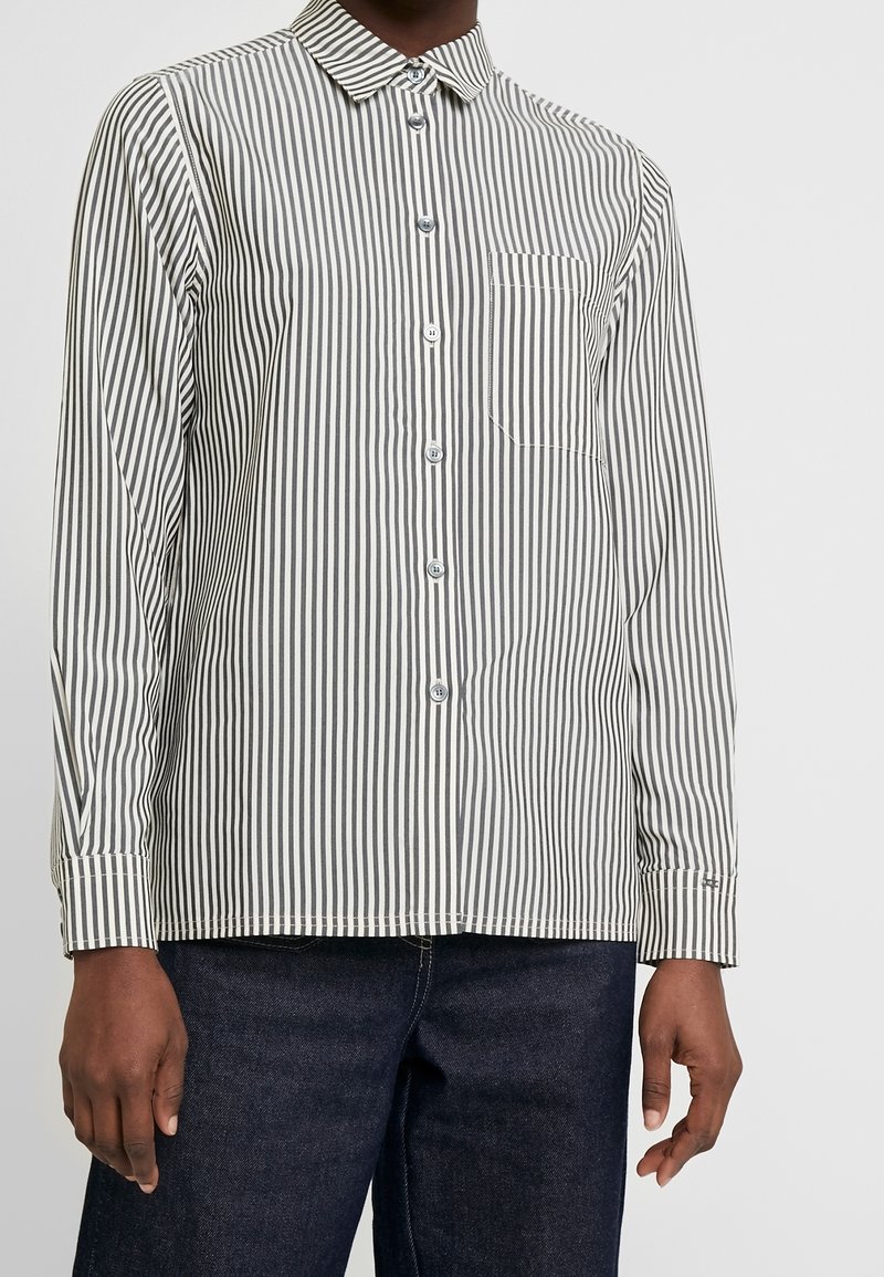 Chemise rayée noire et blanche à boutons avec manches longues et poche avant, portée avec un jean bleu foncé.