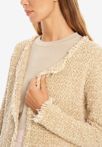 Ljust beige texturerad cardigan med fransar i kanten. Bärs över en krämfärgad topp, och visar en avslappnad passform och en öppen frontdesign. Mjuk material synligt.