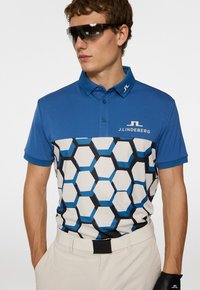 Blå polo t-shirt med geometriskt hexagonmönster i blått, svart och vitt. Har en krage och logotyp på bröstet; kombinerad med beige byxor.