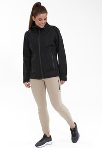 Veste imperméable noire avec un zip sur le devant, deux poches latérales et une capuche, associée à un pantalon fitted beige et des chaussures de sport noires.