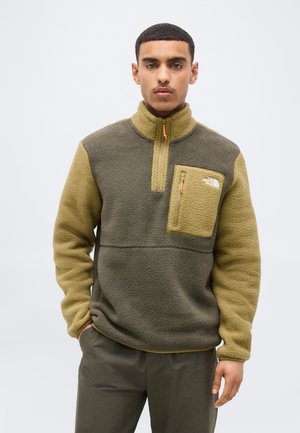 Άνδρας που φοράει μπουφάν από φλις σε ελιά πράσινο και μπεζ χρώμα με μισό φερμουάρ και τσέπη στο στήθος με φερμουάρ, στέκεται μπροστά σε απλό φωτεινό φόντο.