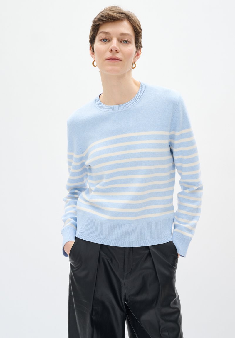 Maglione a righe blu chiaro con strisce orizzontali crema, realizzato in tessuto morbido. Presenta un colletto e dei polsini a coste, abbinato a pantaloni neri.