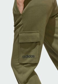 Pantaloni felpati verde oliva con tasca laterale. Realizzati in morbido misto cotone, presentano il logo ricamato in nero "adidas" vicino alla tasca.