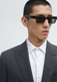 Schwarze Sonnenbrille mit rechteckigem Rahmen, getragen zu einem dunkelgrauen Anzug und weißem Hemd mit Knöpfen. Glatte Texturen und scharfe Linien.