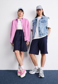 Deux mannequins se tiennent côte à côte. Celui de gauche porte une veste en velours côtelé rose sur un crop top blanc et un short bleu marine. Celui de droite porte un gilet en denim sur une chemise blanche et un short bleu marine. Tous deux portent des chaussures décontractées : des baskets roses à gauche, des baskets grises à droite.