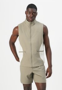 Veste sans manches beige avec un col montant, fermeture éclair sur le devant et panneaux latéraux blancs contrastés. Comprend deux poches zippées à l'avant.
