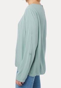 Ljusgrön långärmad blus med rund halsringning och detaljer i silver vid ärmsluten, tillverkad av ett slätt, flytande tyg.