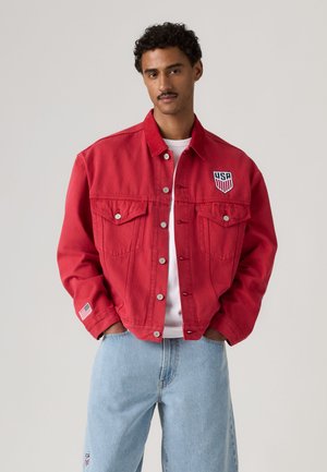 Jeune homme portant une veste rouge boutonnée avec des patchs USA, une chemise blanche et un jean bleu clair sur un fond uni.
