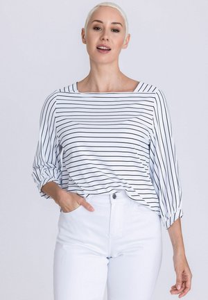Woman with short blonde hair wearing a white top with black horizontal stripes and white high-waisted pants, hand in pocket.