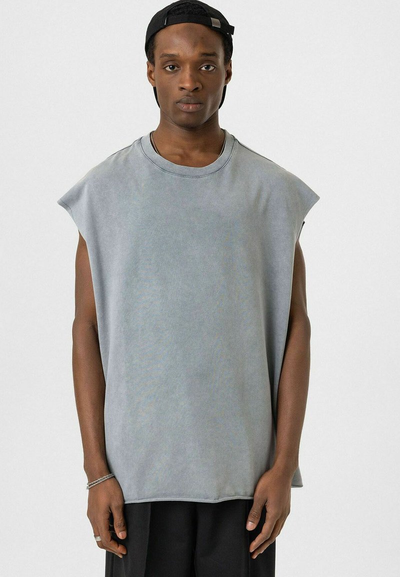 Grijze mouwloze T-shirt gemaakt van zachte stof. Heeft een ronde hals en een losse pasvorm met ruwe randen. Draagt in combinatie met zwarte broeken en een pet.