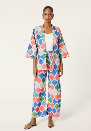 Mujer con chaqueta holgada de estampado multicolor a juego y pantalones anchos, con top blanco y sandalias marrones, de pie frente a un fondo liso.