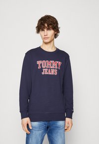 Sudadera azul marino de algodón, con "TOMMY JEANS" en letras rojas en negrita, puños acanalados y un diseño de cuello redondo. Combinada con jeans de color azul claro.