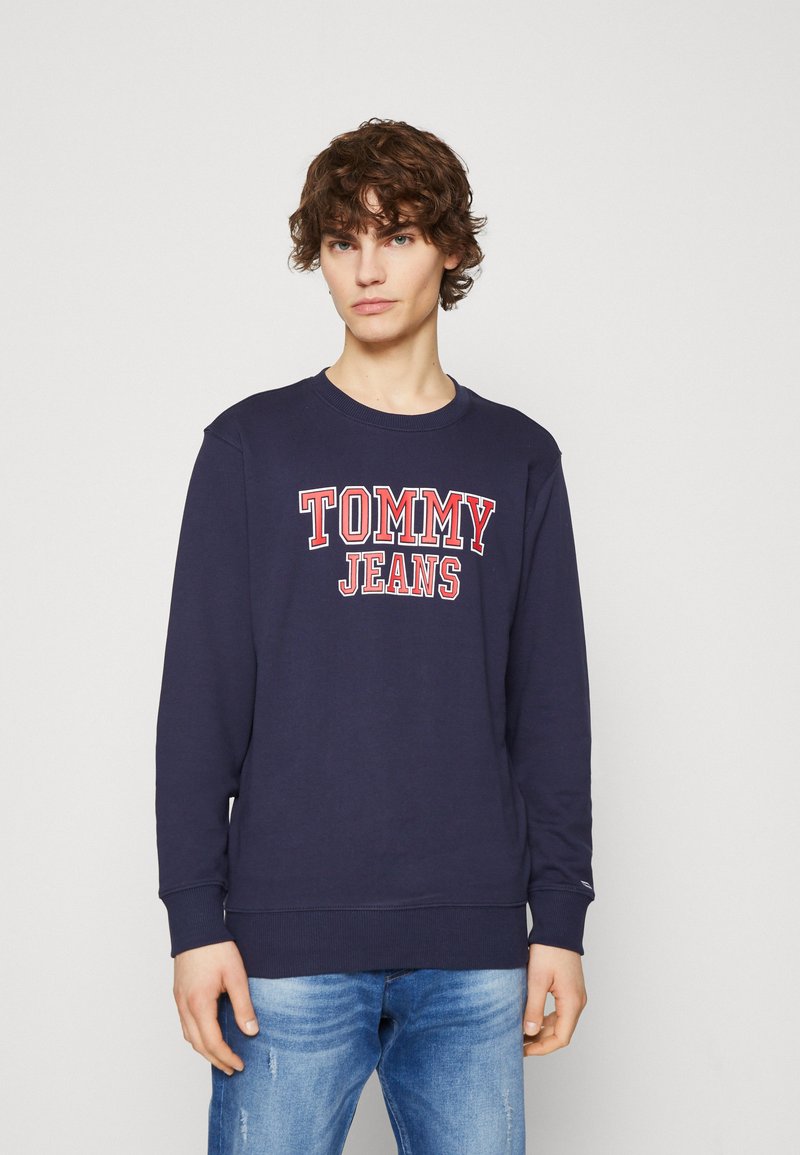 Sudadera azul marino de algodón, con "TOMMY JEANS" en letras rojas en negrita, puños acanalados y un diseño de cuello redondo. Combinada con jeans de color azul claro.