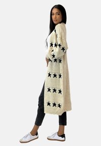 Larga chaqueta de punto crema con patrones de estrellas negras, textura de punto grueso, diseño de frente abierto, combinada con jeans oscuros y zapatillas blancas.