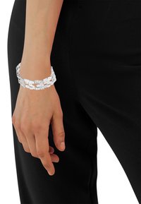 Bracciale in argento con forme geometriche intrecciate e una texture liscia, indossato su un polso su uno sfondo scuro.