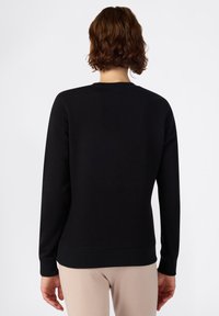 Sudadera negra con mangas largas, cuello redondo y puños acanalados. Textura suave y dobladillo ajustado. Llevada sobre pantalones beige en un fondo neutro.