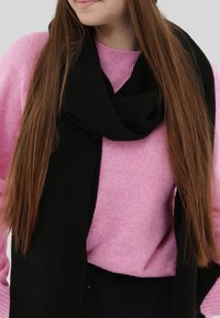 Sciarpa nera con trama a rilievo avvolta intorno al collo, in contrasto con un maglione rosa. La sciarpa presenta un motivo intrecciato spesso, materiale morbido e cade morbida.