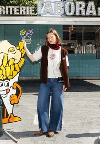 Une femme portant un gilet en polaire marron sur un t-shirt blanc, un jean large bleu et des sabots à imprimé léopard se tient à côté d'un présentoir de frites de style cartoon.