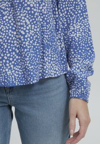 Blusa blu a fantasia con macchie bianche, maniche lunghe con polsini arricciati, abbinata a jeans di denim blu. Tessuto di cotone morbido.