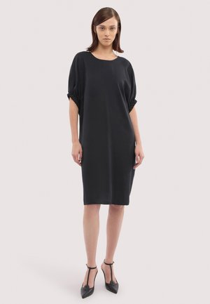 Kobieta stojąca w luźnej czarnej sukience do kolan z marszczonymi krótkimi rękawami oraz czarnych, spiczastych butach na wysokim obcasie z paskiem w kształcie litery T.
