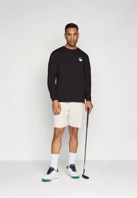 Fekete, hosszú ujjú póló logóval, fehér rövidnadrág és golfcipő. Sportos fazon, kontrasztos részletek és egy golfütő a kézben.