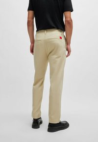 Beige trousers with a tailored fit, made from a smooth fabric. Features rear pockets and a red brand label. Worn with black shoes.