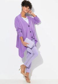 Cardigan oversized color lavanda con bottoni neri, abbinato a pantaloni lavanda con una striscia argentata e tacchi nudi, mentre tiene una pochette trapuntata.