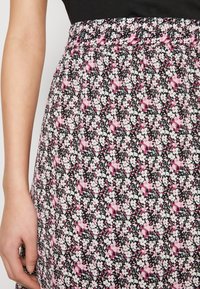 Jupe noire à motif floral avec des fleurs roses et blanches, ceinture élastique et tissu léger, montrée de la taille vers le bas avec une main.