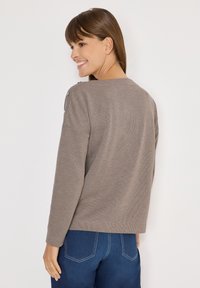 Hellbrauner langärmeliger Pullover mit weitem Ausschnitt und Schulterknöpfen, kombiniert mit blauen Hosen mit kontrastierenden Nähten.