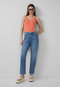 Koraalroze tanktop, lichtblauwe high-waisted jeans en beige plateausandalen. Model draagt oversized zonnebril, neutrale achtergrond.
