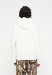 Persona con capelli corti ricci scuri che indossa un felpa bianca semplice e pantaloni a motivo mimetico, vista da dietro su uno sfondo bianco.
