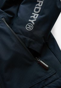 Giacca navale con una texture a coste, dotata di chiusura con zipper e logo "SUPERDRY" in argento. Include un piccolo laccio per un facile accesso.