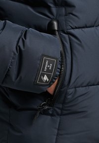 Ręka w kieszeni granatowej puchowej kurtki Superdry, widoczny zamek błyskawiczny oraz czarna naszywka z logo i tekstem na rękawie.