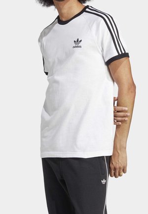 Person trägt ein weißes Adidas-T-Shirt mit schwarzen Streifen auf den Ärmeln und schwarze Adidas-Sweatpants mit weißen Streifen an den Seiten.