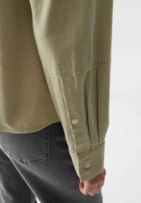 Nahaufnahme eines Arms, der ein beiges Cordhemd mit geknöpften Manschetten über schwarzen Jeans trägt.