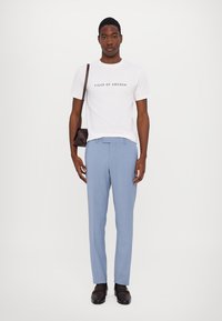 Weißes T-Shirt mit dem Text "TIGER OF SWEDEN", hellblaue maßgeschneiderte Hose und dunkle Lederschuhe. Eine schwarze Umhängetasche hängt über der Schulter.