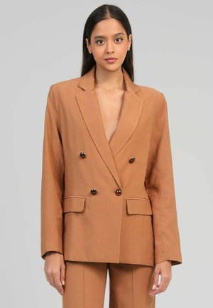 Femme aux longs cheveux foncés portant un blazer marron croisé et un pantalon assorti, se tenant devant un fond gris uni.