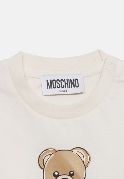 Μπλουζάκι μωρού σε κρεμ χρώμα με ετικέτα Moschino και εκτύπωση με καφέ αρκουδάκι κοντά στον γιακά.