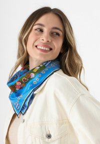 Blauwe zijden sjaal met bloemen- en feestpatronen, met een groene en donkerblauwe rand. Netjes om de nek geknoopt, boven een witte jas.