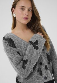 Jauna sieviete ar gaismas brūniem matiem, ģērbta pelēkā pūkainā kardigānā ar melniem putnu motīviem un lielām sudraba auskarām.