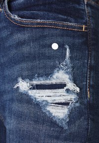 Mörkblå denimjeans har en stor triangelformad reva med fransade kanter och vita horisontella ränder. En liten vit tagg finns ovanför fickan.