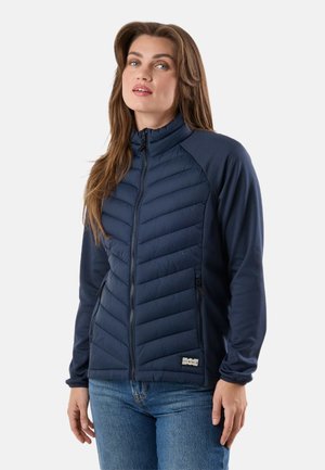 Nainen, joka pukeutunut tummansiniseen tikattuun takkiin, jossa on vetoketjulliset taskut, seisoo yksinkertaisen valkoisen taustan edessä.