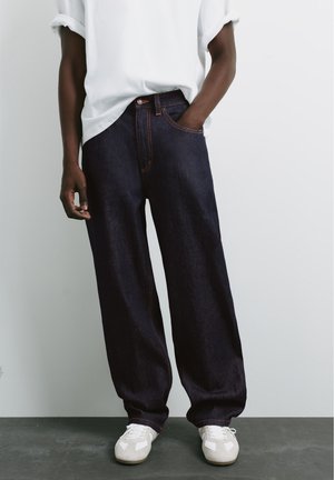 Personne portant un jean bleu foncé ample, une chemise blanche aux manches retroussées et des baskets blanches, debout avec une main dans la poche.